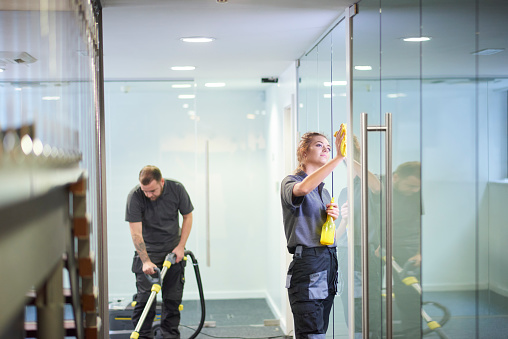 a female cleaning contractor is polishing the glass partition offices whilst In the background a male colleague steam cleans an office carpet in a empty office in between tenants.  .