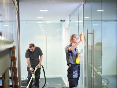 a female cleaning contractor is polishing the glass partition offices whilst In the background a male colleague steam cleans an office carpet in a empty office in between tenants.  .