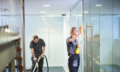 a female cleaning contractor is polishing the glass partition offices whilst In the background a male colleague steam cleans an office carpet in a empty office in between tenants.  .